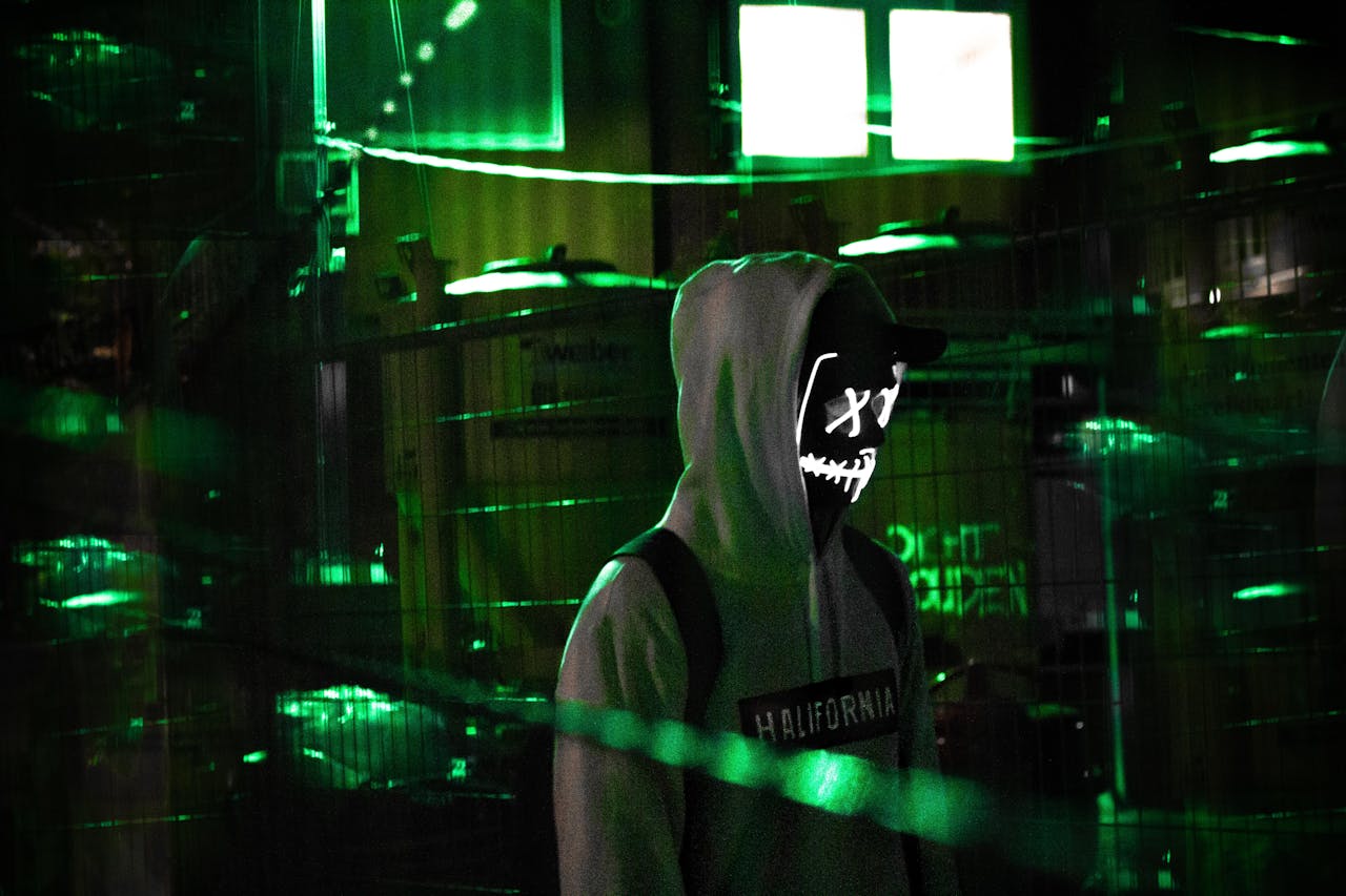 Man in a hoodie with glowing mask amidst neon green lights, evoking mystery and intrigue.